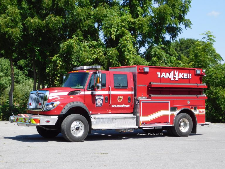 A red fire truck with "TANKER" written on the side is parked on a paved surface, with green trees and blue sky in the background.
