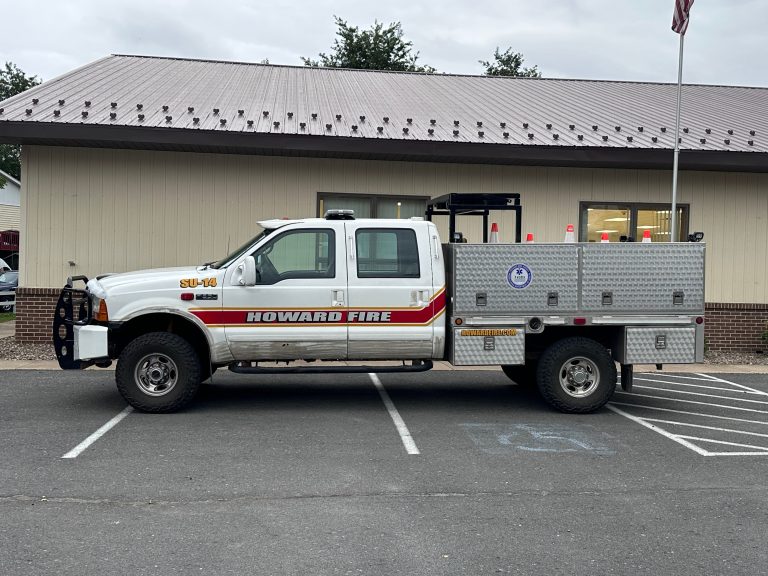 A white Howard Fire Special Unit 14 utility truck with red stripes is parked in a lot, occupying two spaces, including a handicapped spot. The truck has emergency lights, equipment storage, and orange safety cones in the back.