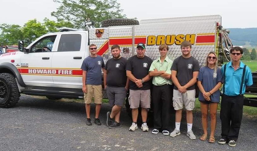 Seven people stand in a row in front of a Howard Fire “Brush” truck, smiling for the camera. The truck is white with red and yellow stripes, parked on a gravel area with fields and trees in the background.