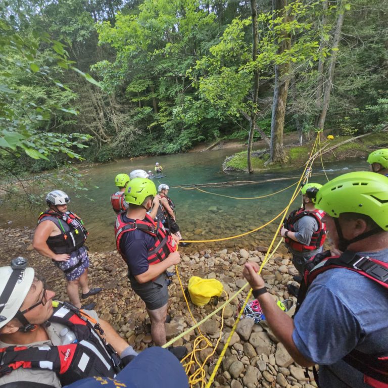 A group from Citizens Hose Company, wearing helmets and life vests, stand on a rocky riverbank holding yellow ropes, preparing for joint water rescue training by a tree-lined river.