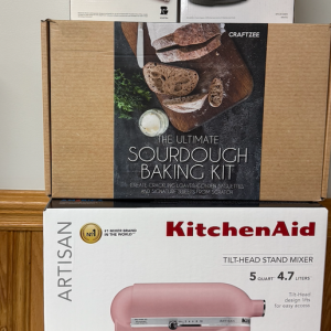 RAFFLE #568 features a Pink 5Qt KitchenAid Stand Mixer with attachments and a Sourdough Starter Kit, all packaged and displayed against wood paneling—an ideal prize for any kitchen enthusiast.