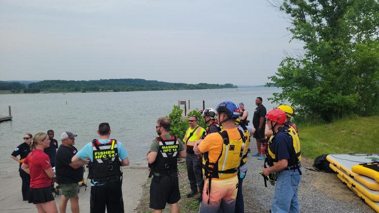 A group of people in safety gear and life jackets gathers near a lakeshore, listening to instructions for rescues. Some wear helmets. Inflatable rescue boats and trees are nearby; the water and distant hills reflect strong community support.