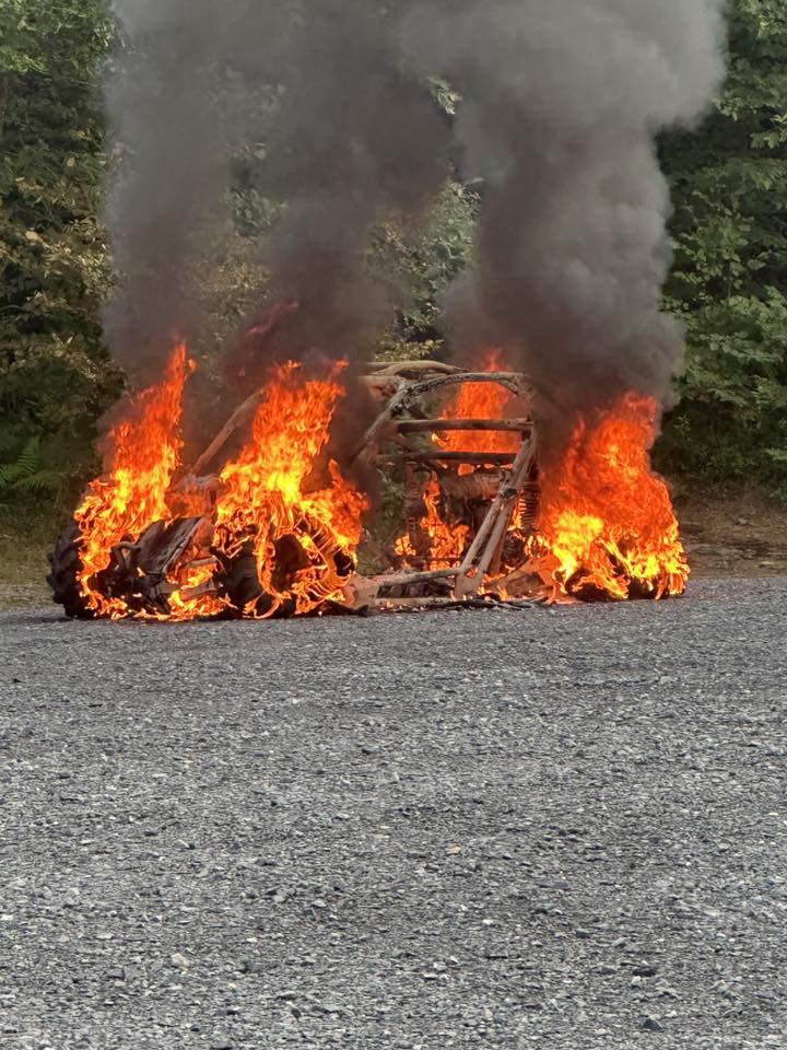 A vehicle engulfed in bright orange flames and thick black smoke sits on a gravel surface, surrounded by trees in the background. The intense Curtin Township Incident has left only the frame of the vehicle visible.