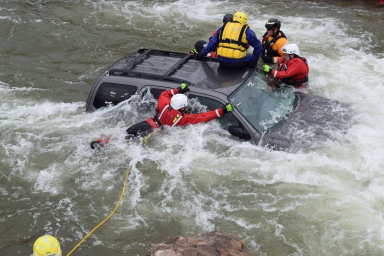 Four rescue workers from the Howard Volunteer Fire Company in helmets and wetsuits rescue someone from a partially submerged SUV in fast-moving water. Two are on top of the vehicle, while two others hold its side. A yellow rope is attached.