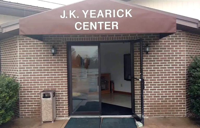 Entrance to the J.K. Yearick Center, featuring a glass door, a maroon awning with the center’s name in white letters, brick exterior walls, a trash can, and bushes on either side—an inviting spot to rent space for your next event.
