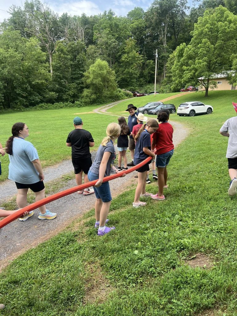 A group of children and adults stand outdoors on a grassy area at Junior Grange Camp, holding a long red tube together. Trees, parked cars, and a gravel path are visible in the background as they practice teamwork.