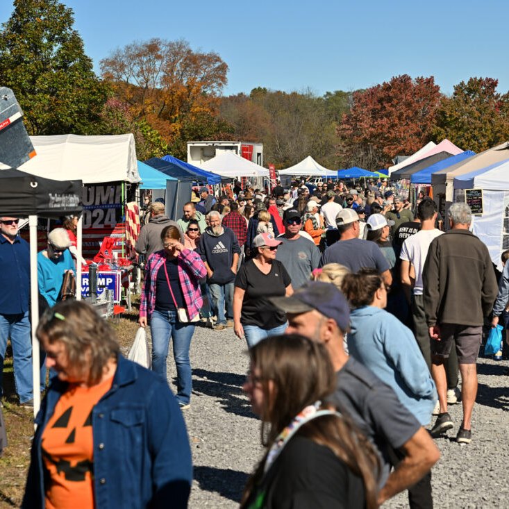 A large crowd of people walks through an outdoor market at a lively Fall Festival, with vendor tents on both sides and autumn trees in the background.