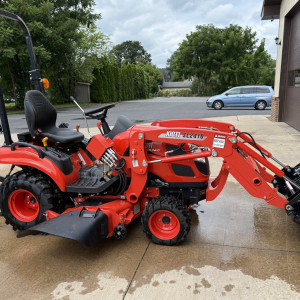 A bright red Kioti CS2210HB Sub-Compact Tractor with loader and 60" mower deck (Raffle #???) is parked on a wet driveway beside a building, with trees and a gray car in the background.