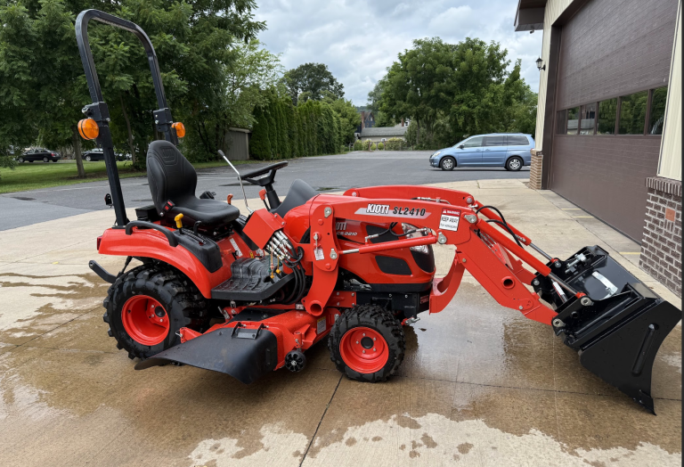 A bright red Kioti CS2210HB Sub-Compact Tractor with loader and 60" mower deck (Raffle #???) is parked on a wet driveway beside a building, with trees and a gray car in the background.