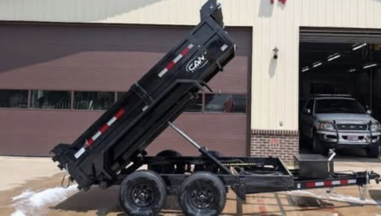 A black double-axle dump trailer with reflective stripes is parked outside a garage. Inside, a Raffle #??? - Kioti CS2210HB Sub-Compact Tractor W/ Loader and 60" mower deck is partially visible.