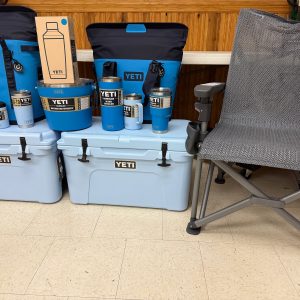 The Raffle #??? - Blue YETI Package, featuring coolers, drinkware, and bags in blue, is displayed next to a gray mesh camp chair on a tiled floor against a wooden wall—a great choice for your next raffle prize.