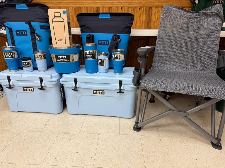 The Raffle #??? - Blue YETI Package, featuring coolers, drinkware, and bags in blue, is displayed next to a gray mesh camp chair on a tiled floor against a wooden wall—a great choice for your next raffle prize.