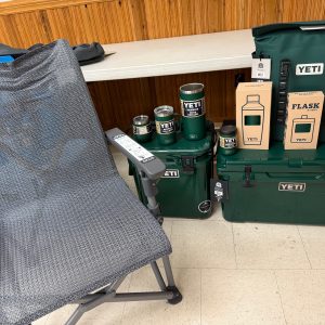 A gray mesh camping chair sits beside a table showcasing the Raffle #598 - Black Forest Green YETI Package—coolers, tumblers, mugs, water bottles, and two boxed flasks—displayed indoors on a tiled floor.
