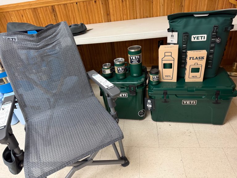 A gray mesh camping chair sits beside a table showcasing the Raffle #598 - Black Forest Green YETI Package—coolers, tumblers, mugs, water bottles, and two boxed flasks—displayed indoors on a tiled floor.