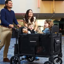 A man and woman smile while pushing the Raffle#609 - 4 Seater WonderFold W4 Luxe Pro Quad Stroller Wagon with Snack Tray and Cargo Net, carrying three children inside, in a bright, modern indoor space.