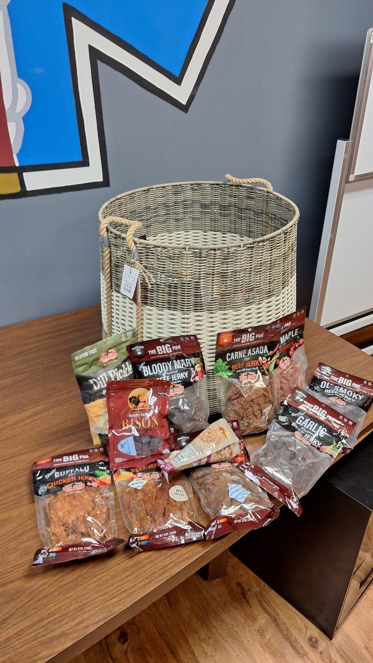 A wicker basket, Raffle #651 - Jumbo Basket Full of Craft Jerky, Exotic Jerky, Meat Sticks, and Flavored Popcorn ($1,500 value), sits on a wooden table with colorful wall art and office furniture in the background.