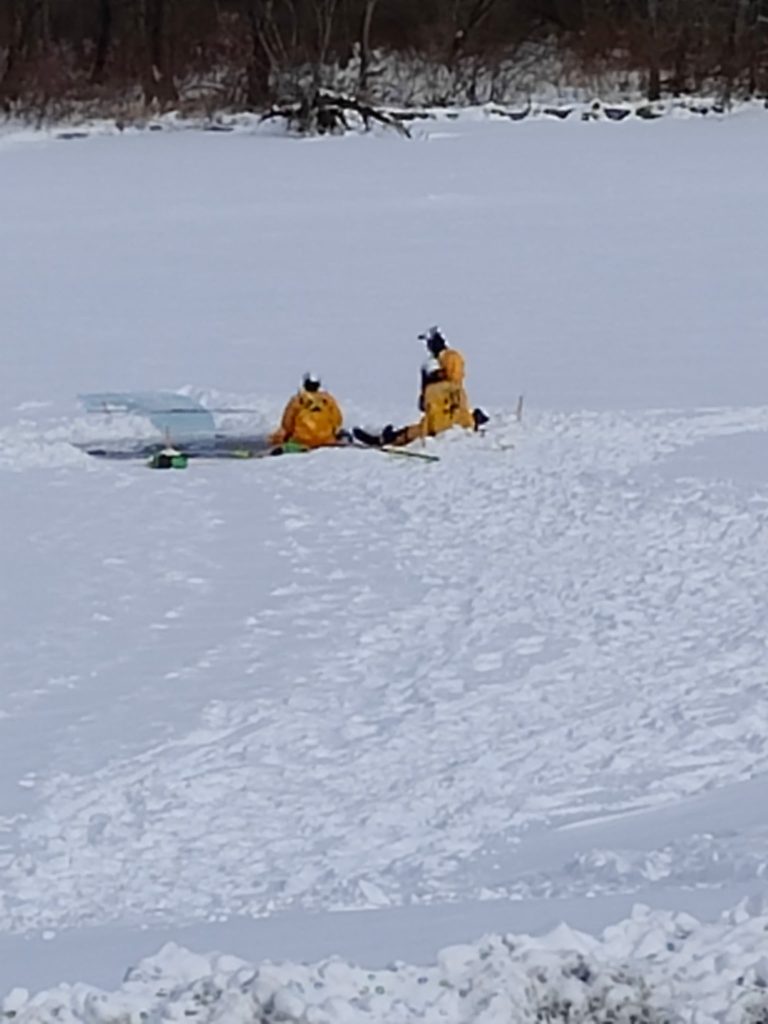 Two people in yellow rescue suits kneel on a snowy, ice-covered surface near broken ice, demonstrating water rescue training as tracks lead to a wooded area in the background.
