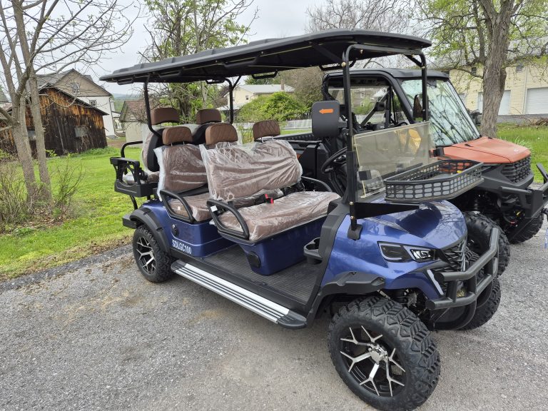 A Raffle #727 Blue 6 Seater 60V Electric Golf Cart with plastic-covered seats is parked outdoors beside a utility vehicle. A 7x14 utility trailer, tax, and tags are included; trees, grass, and buildings are visible in the background.