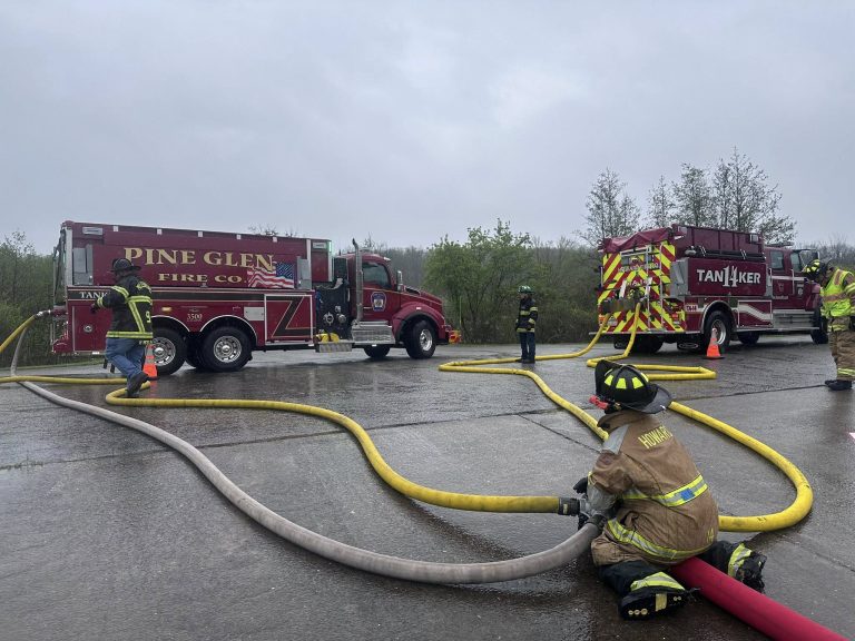 Firefighters in full gear manage hoses connected to two red Pine Glen Fire Company tanker trucks during a firefighter training update on a wet, overcast day. The hoses are spread out on the wet pavement, with trees in the background.