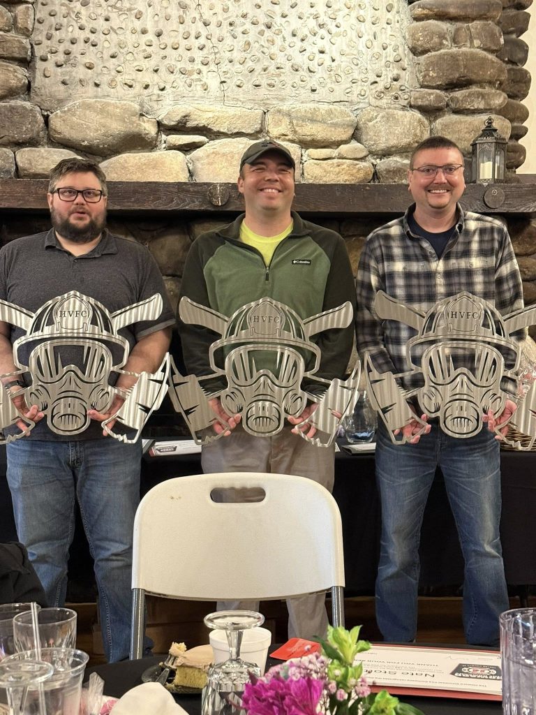Three men stand side by side indoors, each holding large, metallic awards shaped like stylized gas masks. They are smiling at a company banquet in front of a stone fireplace, with a white chair and tables in the foreground.